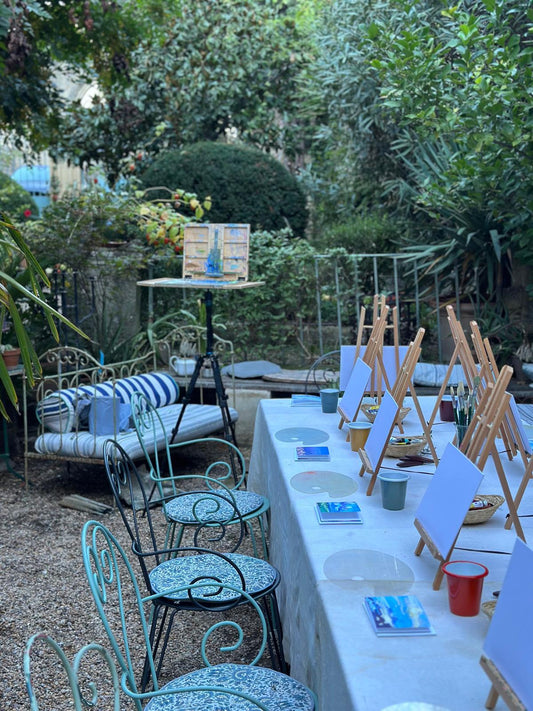 Painting workshop table set with easels and blank canvases in a leafy garden, Lucy Kent's easel behind